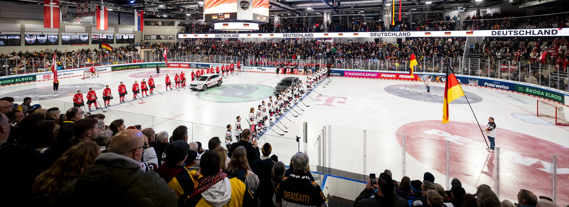 Während der Nationalhymnen stehen sich die Eishockey-Mannschaften Österreich und Deutschland auf dem Eis gegenüber. Zwei Škoda-Fahrzeuge, ein weißer und ein grauer, befinden sich zentral auf der Eisfläche.