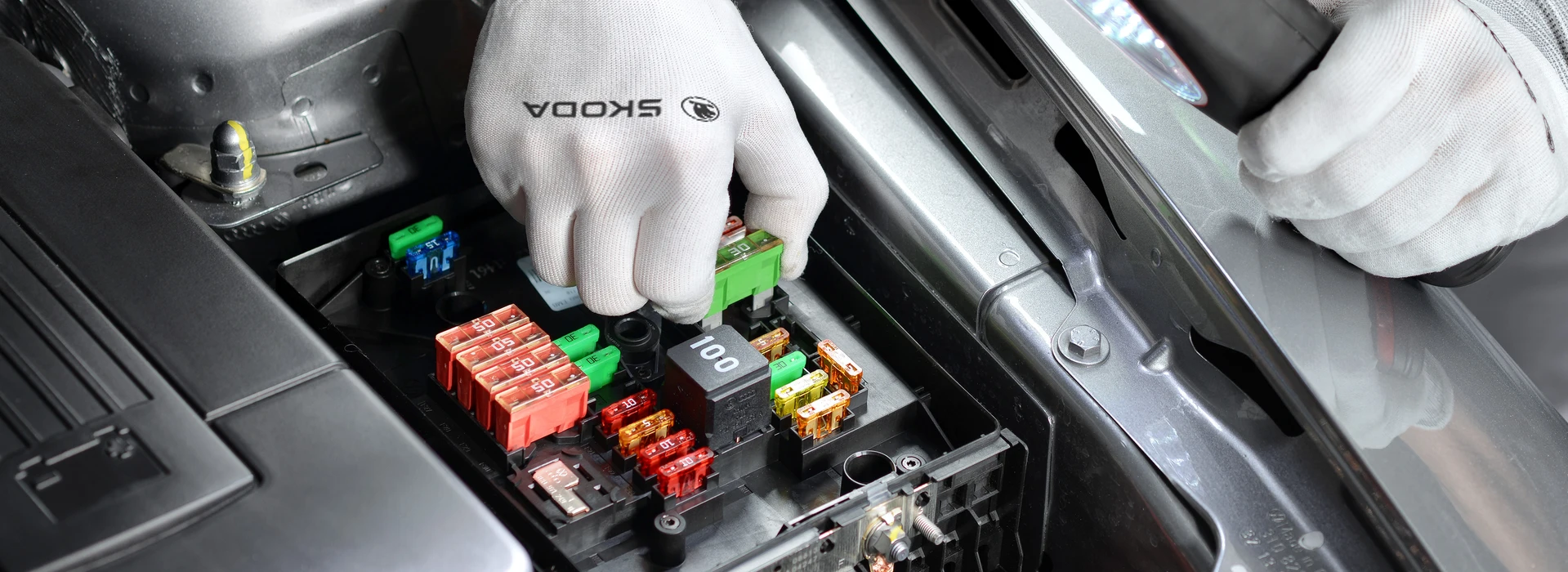 Nahaufnahme des Sicherungskastens eines Škoda-Fahrzeugs. Eine Person mit Handschuhen arbeitet daran und prüft die verschiedenen farbigen Sicherungen.