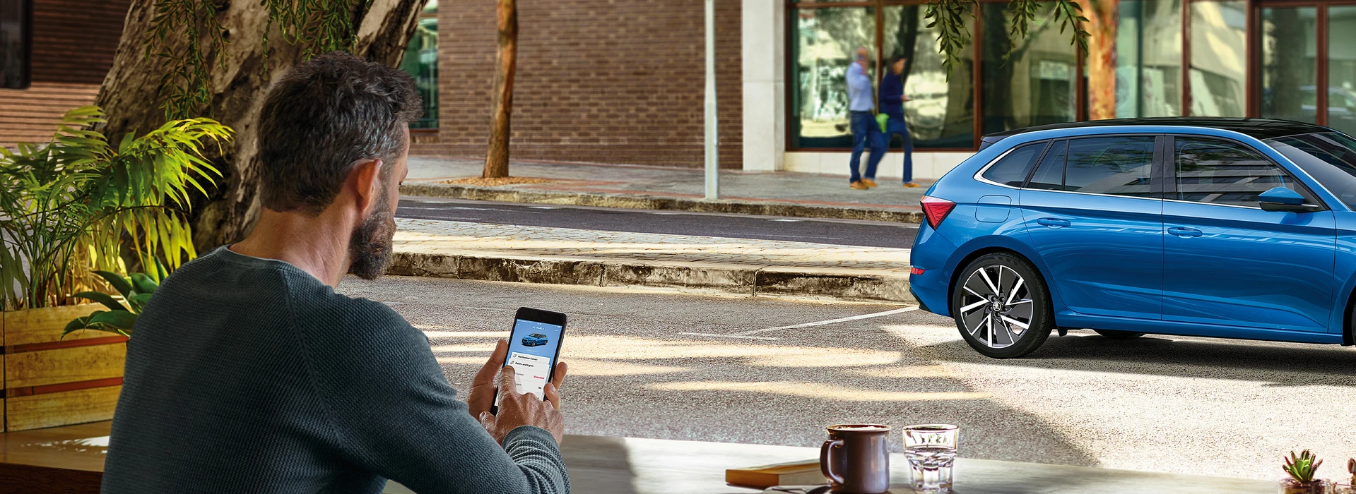 Mann schaut auf Smartphone mit Škoda-Broschüren, im Hintergrund steht ein blauer Kompaktwagen.