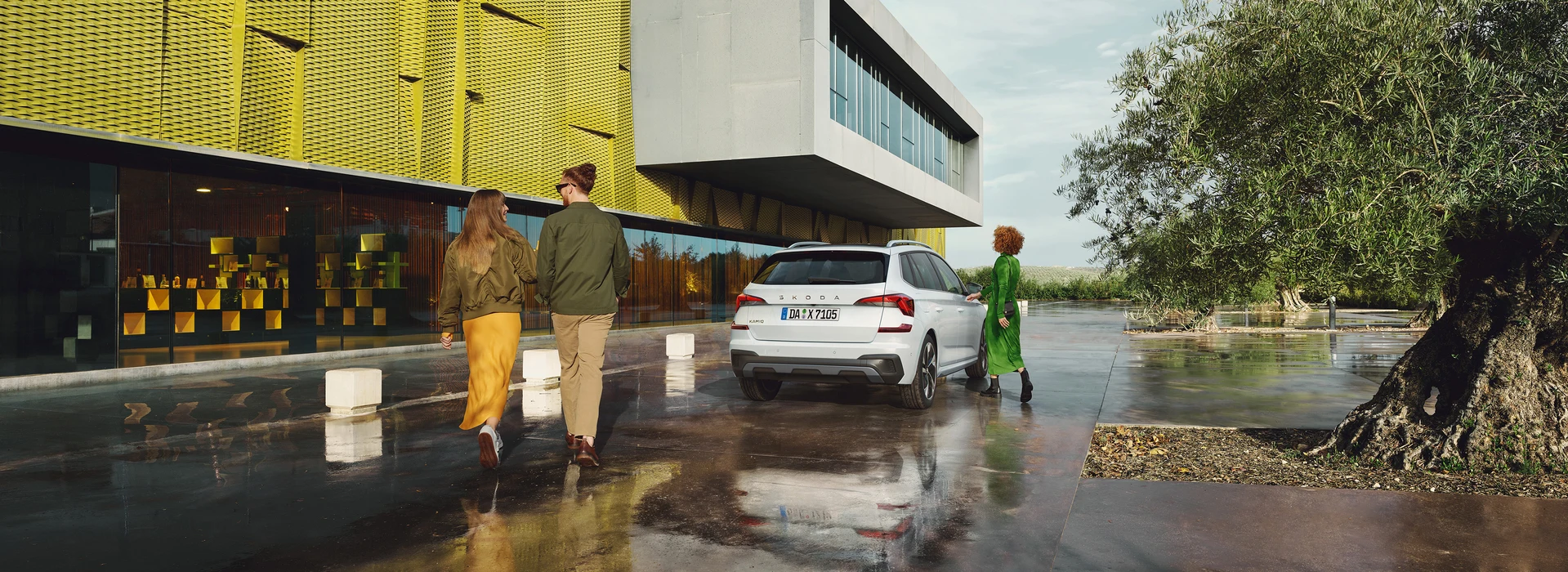 Ein weißer Škoda Kamiq steht auf einer glänzenden Oberfläche, die durch Nässe reflektiert. Im Hintergrund ist ein modernes Gebäude mit gelber Fassade und großen Glasfenstern sichtbar. Drei Personen bewegen sich um das Fahrzeug.
