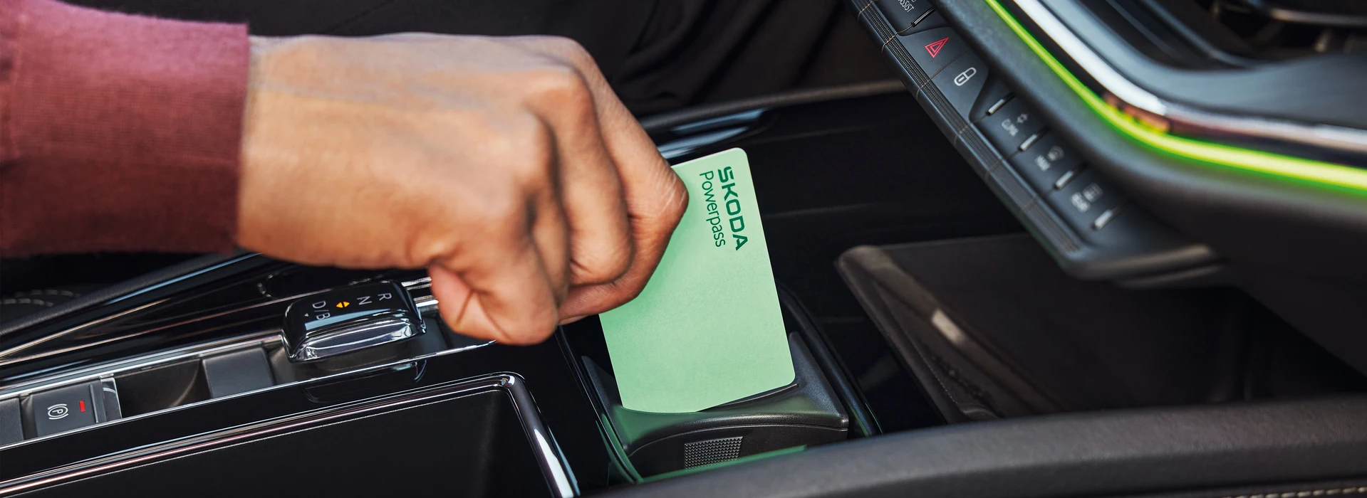 Nahaufnahme einer Hand, die eine grüne ŠKODA Powerpass Ladekarte in das dafür vorgesehene Fach in der Mittelkonsole eines Škoda Fahrzeugs steckt.