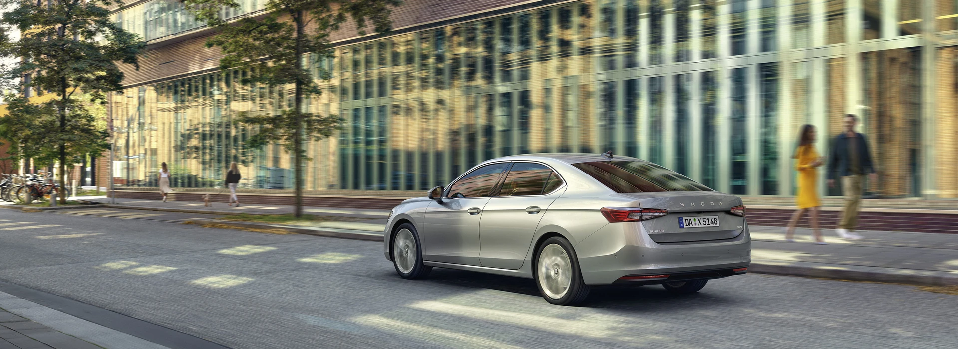 Ein silberner Škoda Superb fährt eine städtische Straße entlang, mit modernen Gebäuden und Bäumen im Hintergrund, während unscharfe Fußgänger auf dem Gehweg zu sehen sind.
