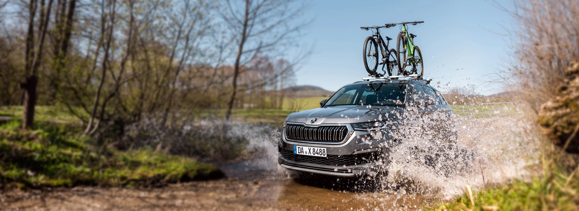 Grauer Škoda SUV mit zwei Fahrrädern auf dem Dach fährt durch Wasser in einer Waldlandschaft.