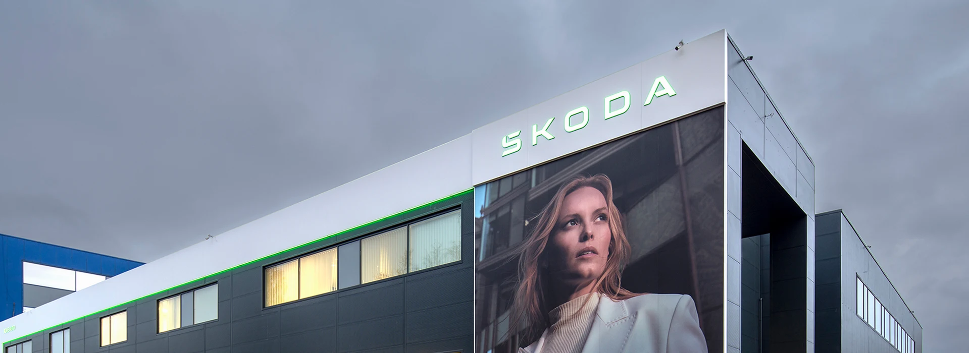 Ein modernes Škoda-Gebäude mit beleuchtetem Logo, großen Glasfronten und einer stilvollen Werbefläche mit einer Frau in elegantem Outfit, vor einem dunklen Himmel