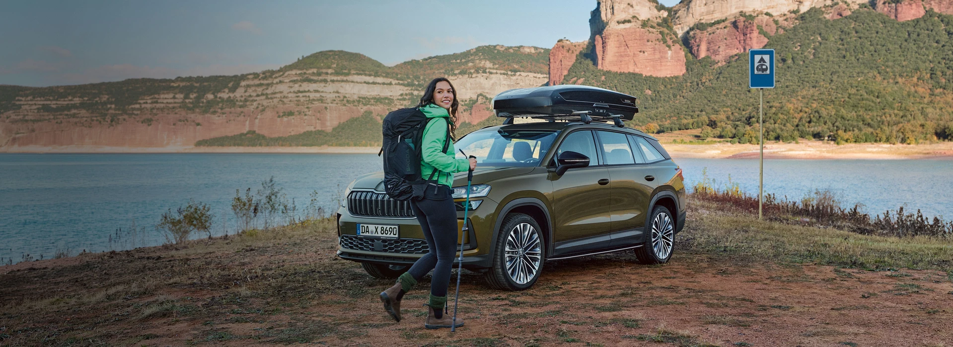 Wanderin mit Rucksack vor einem Škoda Kodiaq mit Dachbox an einem See in bergiger Landschaft.