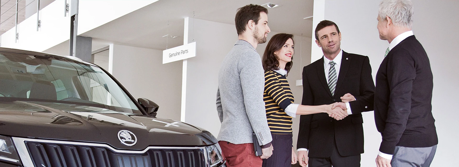 In einem Autohaus stehen vier Personen beisammen, während zwei von ihnen sich die Hand geben. Ein schwarzer Škoda ist im Vordergrund ausgestellt, und ein Schild mit „Genuine Parts“ ist an der Wand befestigt.