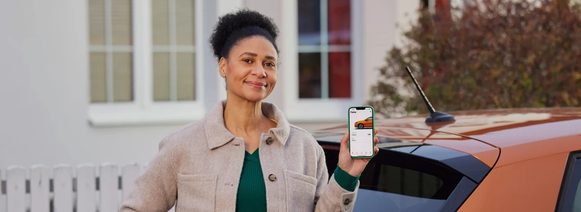 Lächelnde Frau vor Fahrzeug hält Smartphone in die Kamera, auf dem die MyŠkoda App geöffnet ist.