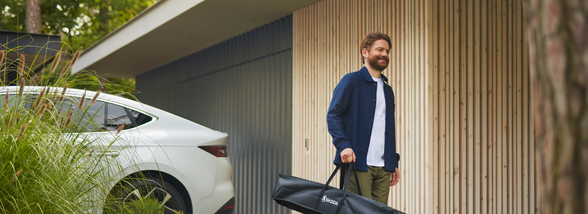 Mann mit Bart trägt schwarze Škoda-Tasche vor modernem Holzhaus mit weißem Auto im Hintergrund.