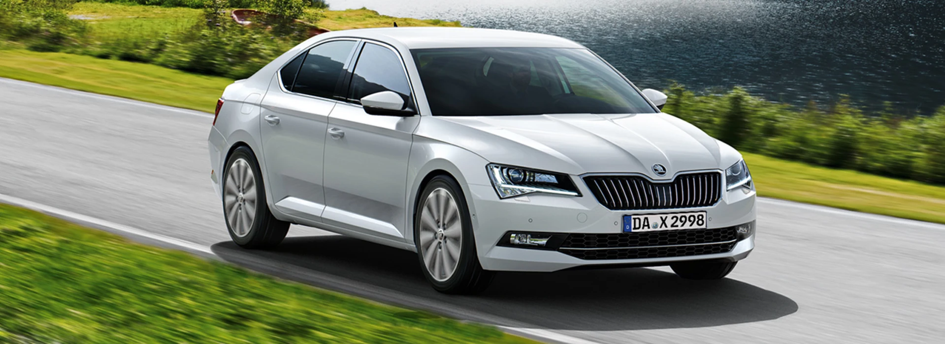 Ein silberner Škoda fährt auf einer Straße neben einem See, im Hintergrund sind grüne Hügel und Bäume.