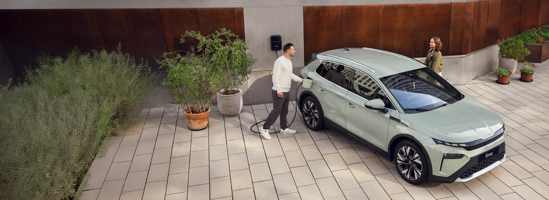 Ein Mann lädt einen hellgrünen Škoda Elroq an einer Wallbox auf, während eine Frau lächelnd danebensteht. Die Szene spielt sich in einem modernen Innenhof mit Pflanzen und minimalistischer Architektur ab.