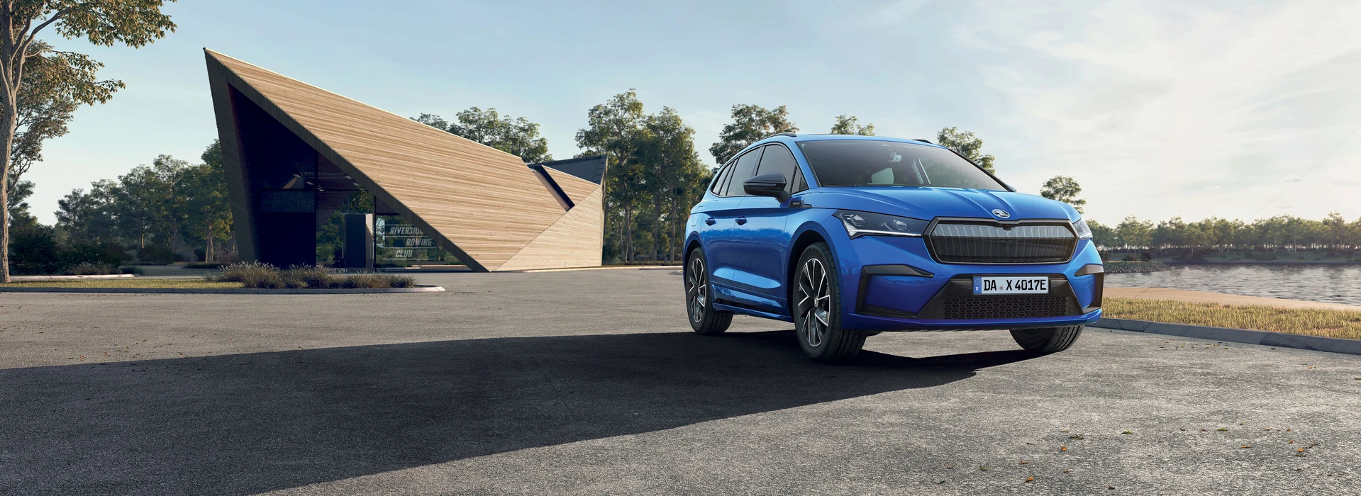 Blauer Škoda Enyaq steht auf einem Platz vor einem modernen Holzbau im Grünen.