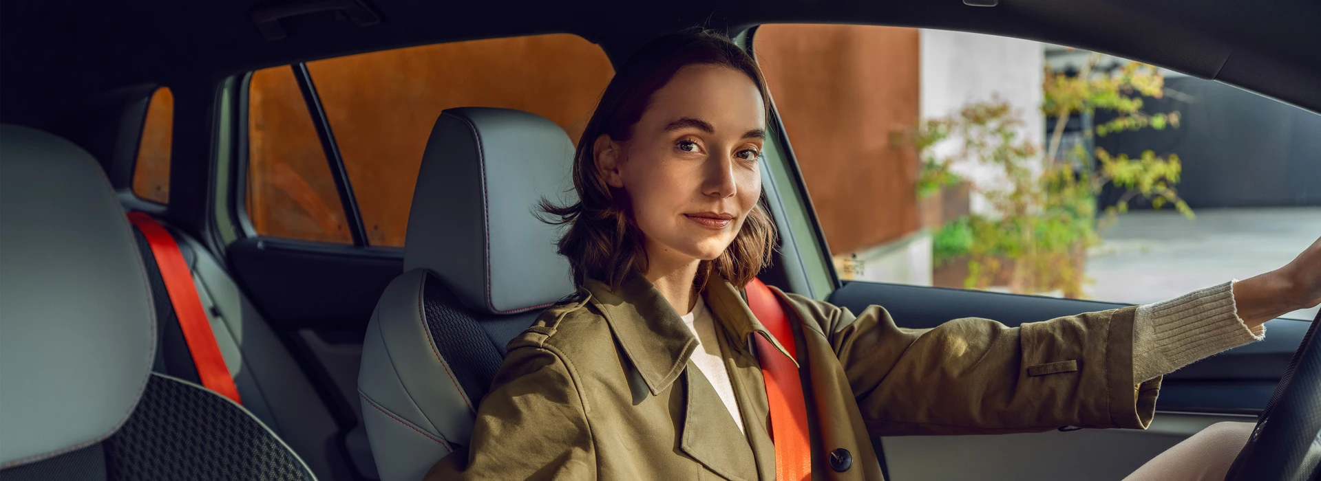 Eine junge Frau mit braunen Haaren und einem grünen Mantel sitzt am Steuer eines Škoda mit orangem Gurt und blickt nach vorn.