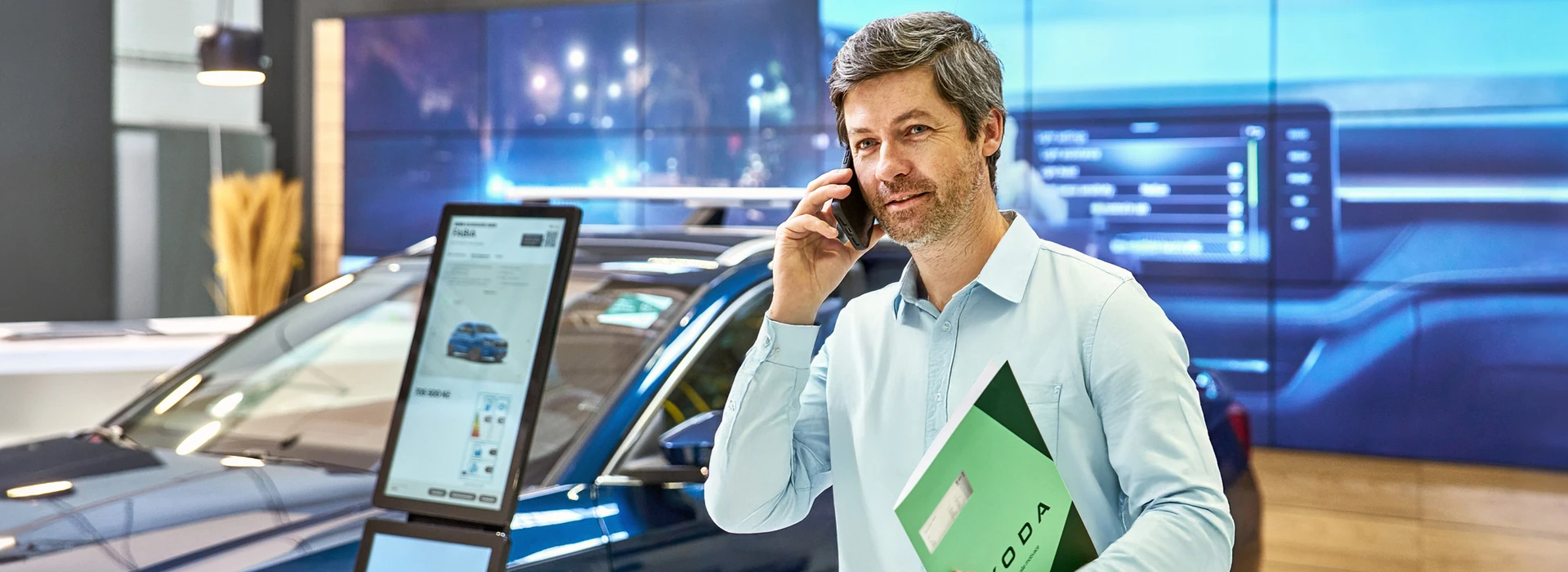 Škoda-Verkäufer mit grauem Haar telefoniert lächelnd, hält grüne Mappe.