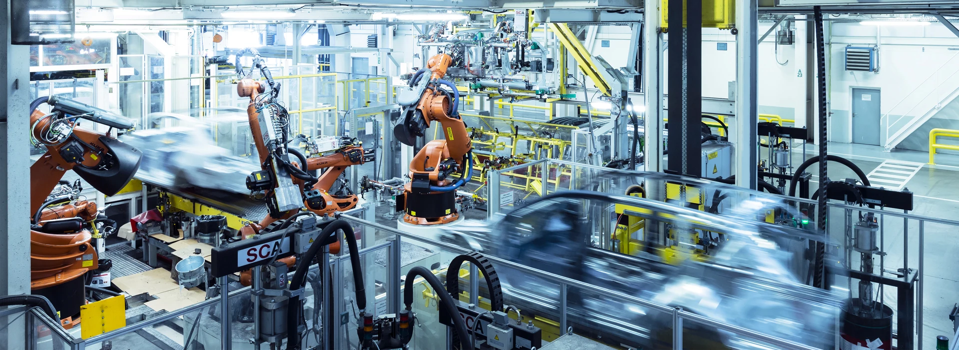 Eine Produktionsstätte mit mehreren orangefarbenen Roboterarmen, die an Fahrzeugkomponenten arbeiten. Förderbänder transportieren Bauteile durch verschiedene Fertigungsabschnitte, während Sicherheitsgeländer die Bereiche strukturieren.