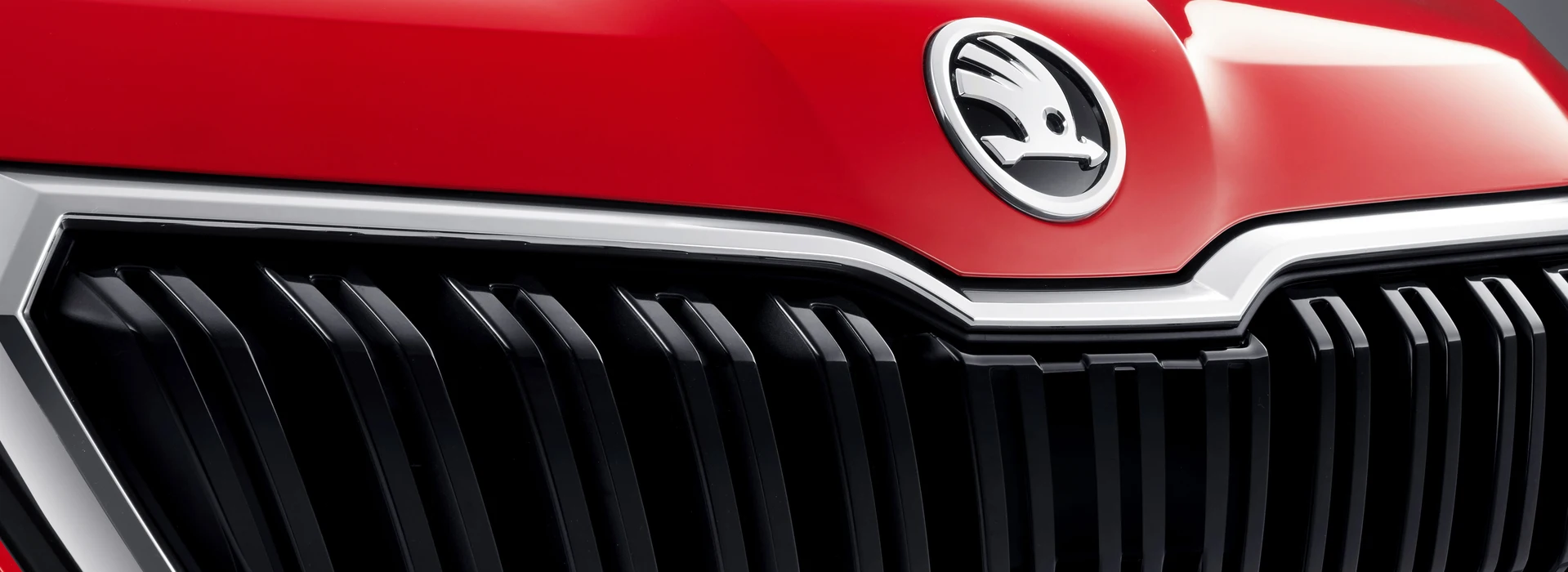 Das Bild zeigt das Frontgrill eines roten Škoda mit vertikalen schwarzen Lamellen, umrandet von Chrom. Oberhalb des Grills prangt das Škoda-Emblem mit dem geflügelten Pfeil.