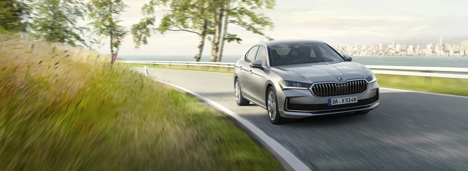 Silberner Škoda Superb fährt dynamisch auf Landstraße entlang einer Küstenstraße.