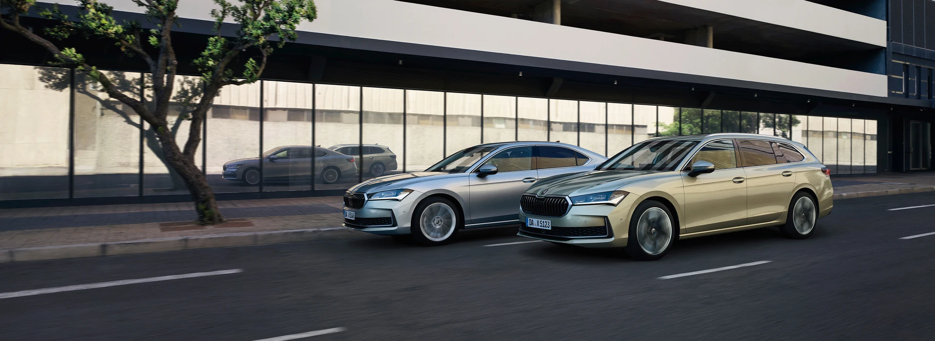 Zwei Škoda Superb Modelle fahren nebeneinander an moderner Glasfassade entlang.
