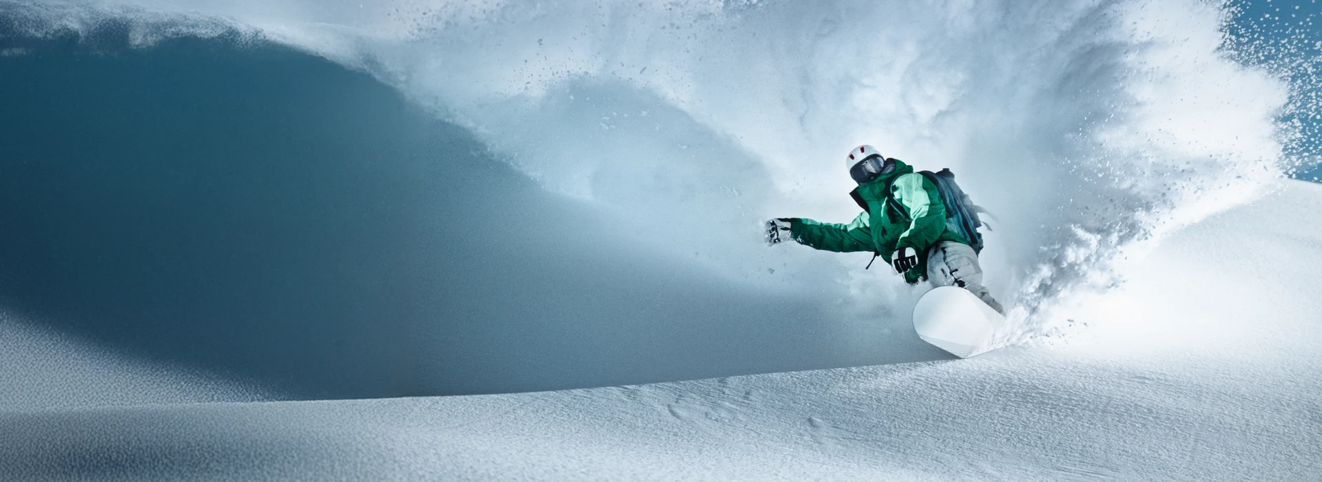 Snowboarder in grüner Jacke fährt dynamisch durch frischen Pulverschnee und wirbelt Schnee auf.