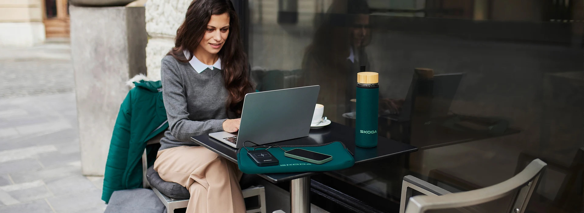 Frau arbeitet im Café am Laptop, mit Škoda-Trinkflasche und Zubehör auf dem Tisch.