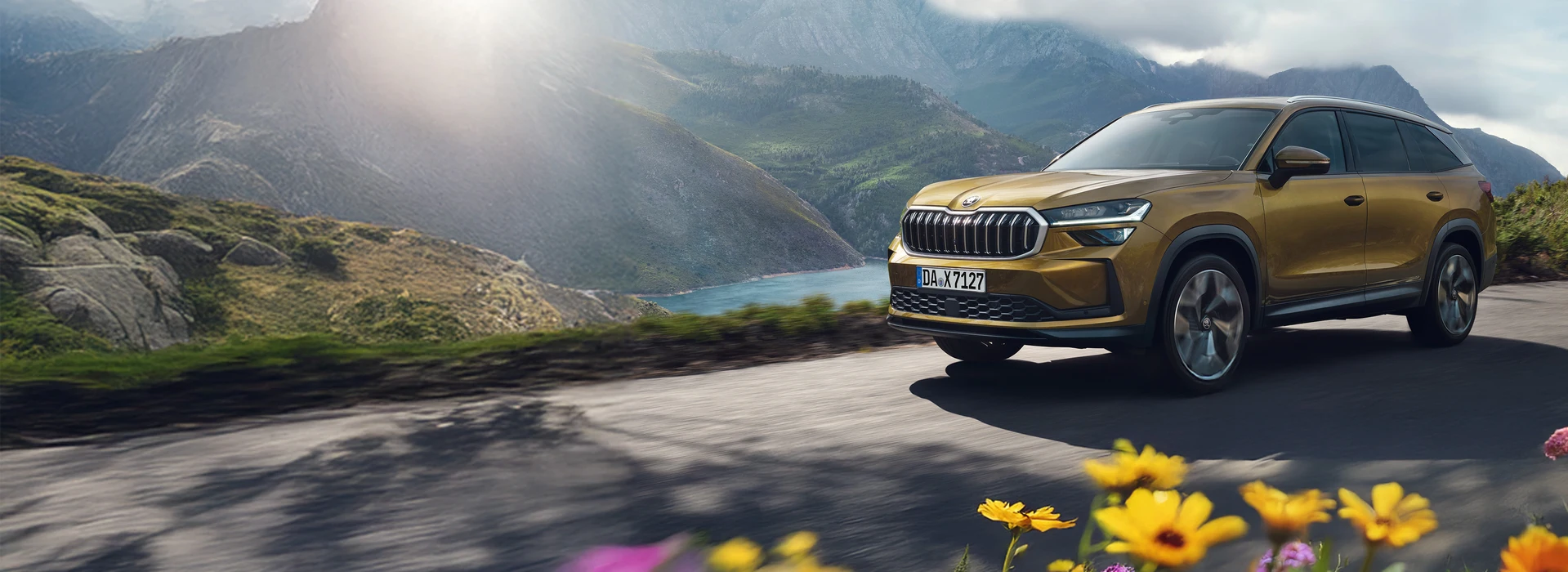 Neuer Škoda Kodiaq in Gold-Bronze Metallic fährt auf Bergstraße am See entlang, sonnige Landschaft im Hintergrund
