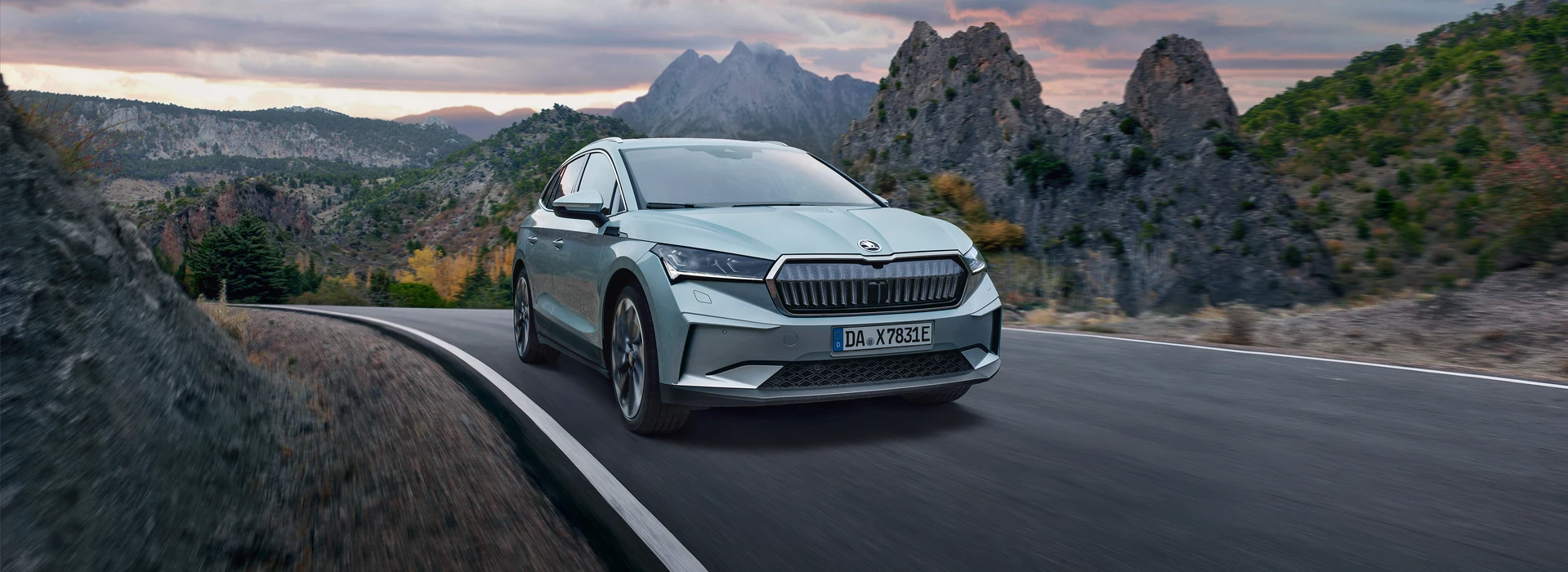 Škoda Enyaq fährt dynamisch auf kurviger Landstraße vor malerischer Bergkulisse.