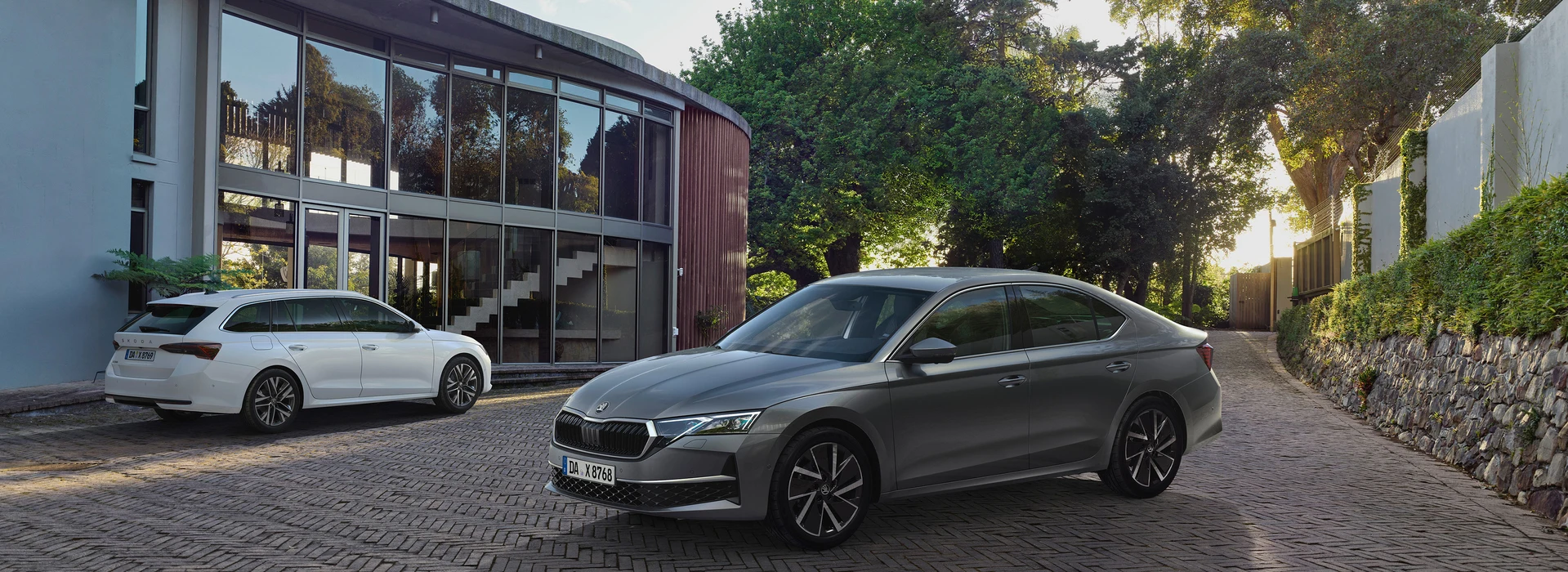 Ein Škoda Octavia und ein Škoda Octavia Combi sind auf einer Auffahrt vor einem Gebäude mit großen Glasfenstern geparkt. Eine Treppe ist durch die Fenster sichtbar, und Bäume sowie Grünflächen umgeben die Szene.