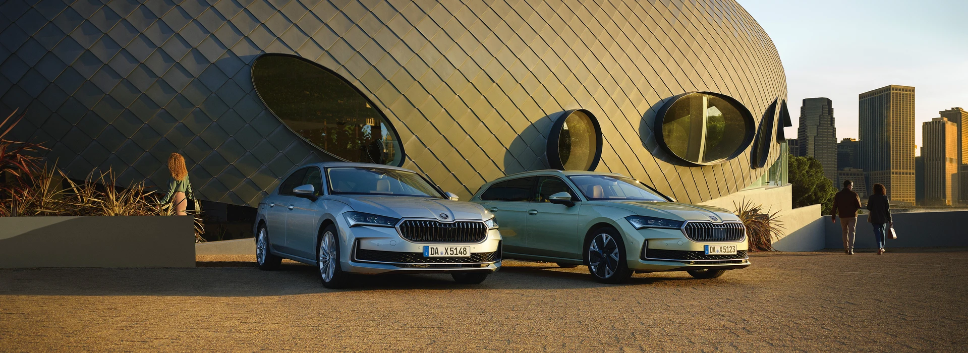 Zwei neue Škoda Superb Modelle (Limousine und Combi) stehen vor einem modernen Gebäude mit einer Stadt-Skyline im Hintergrund.