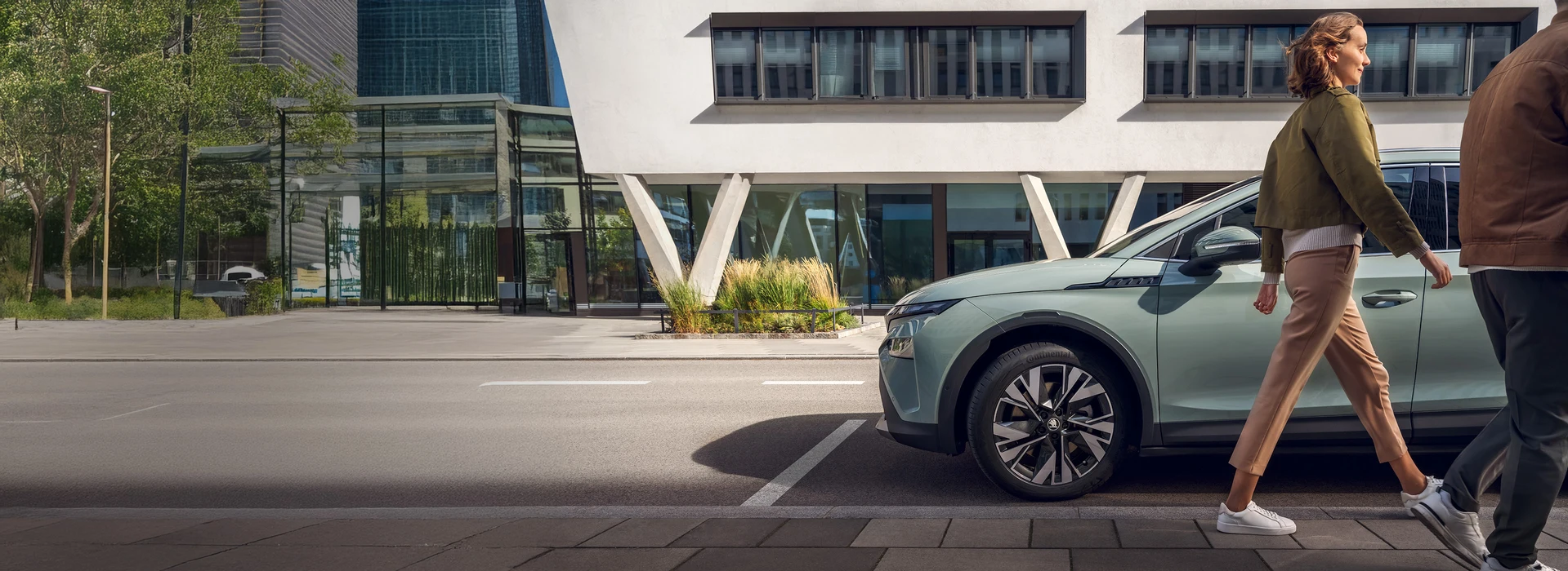 Eine Frau und ein Mann gehen auf dem Gehweg an einem geparkten hellgrünen Škoda Elroq vorbei. Im Hintergrund moderne Architektur mit großen Fenstern und begrüntem Straßenraum.