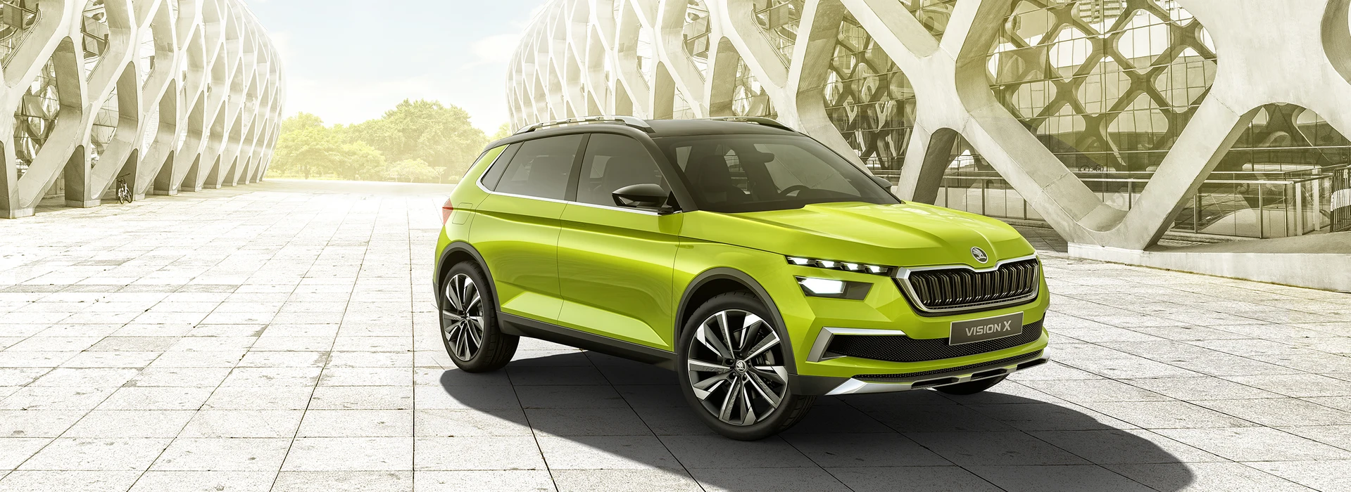 Škoda Vision X Konzeptfahrzeug in leuchtendem Grün, Frontansicht mit beleuchteten LED-Scheinwerfern, markantem Kühlergrill und „VISION X“-Kennzeichen, vor moderner Architektur mit geometrischen Strukturen.