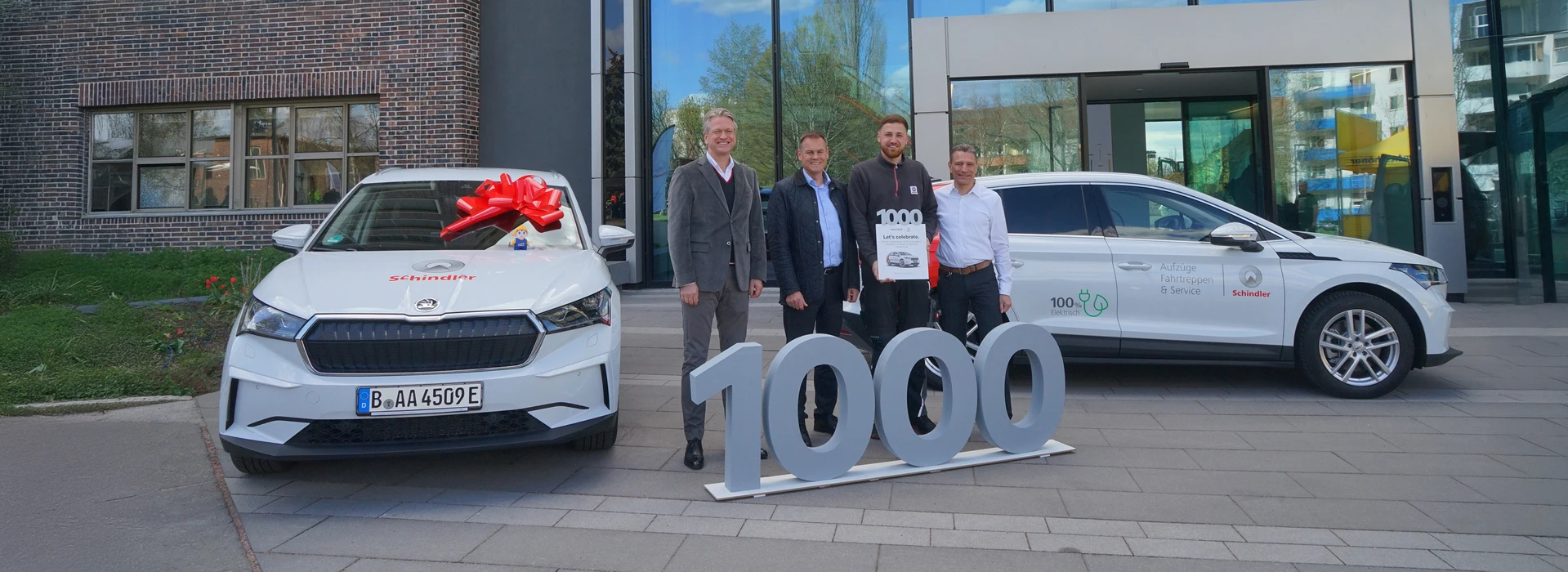 Vier Männer stehen vor zwei Škoda Enyaq, eines mit Schleife, und feiern das 1000. Fahrzeug.