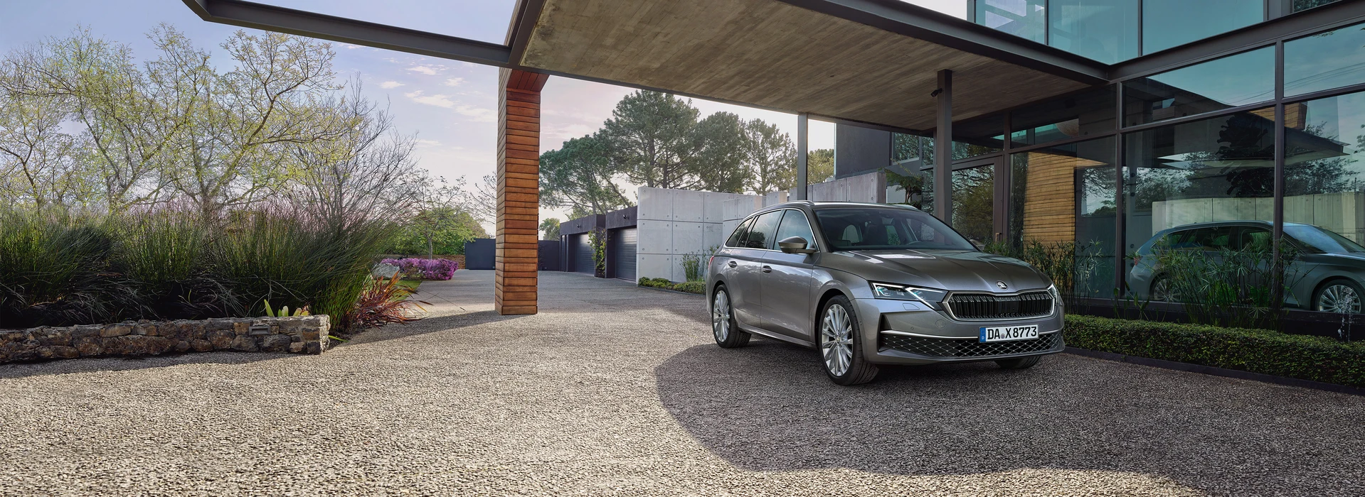 Grauer Škoda Octavia Combi vor moderner Architektur mit Glasfassade und Carport, umgeben von Grünpflanzen.