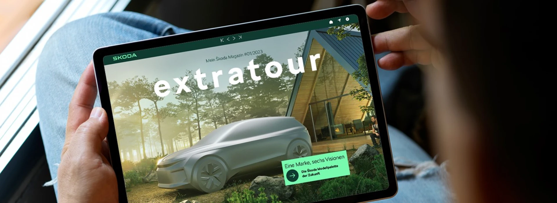 Person hält Tablet mit Škoda Magazin „extratour“ und Konzeptfahrzeug in Waldumgebung.
