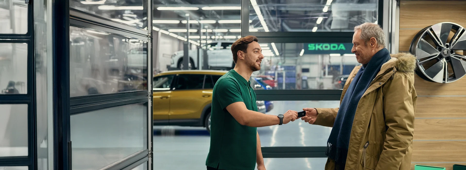 Junger Škoda-Mitarbeiter in grünem Polo übergibt Autoschlüssel lächelnd an älteren Kunden in der Werkstatt.