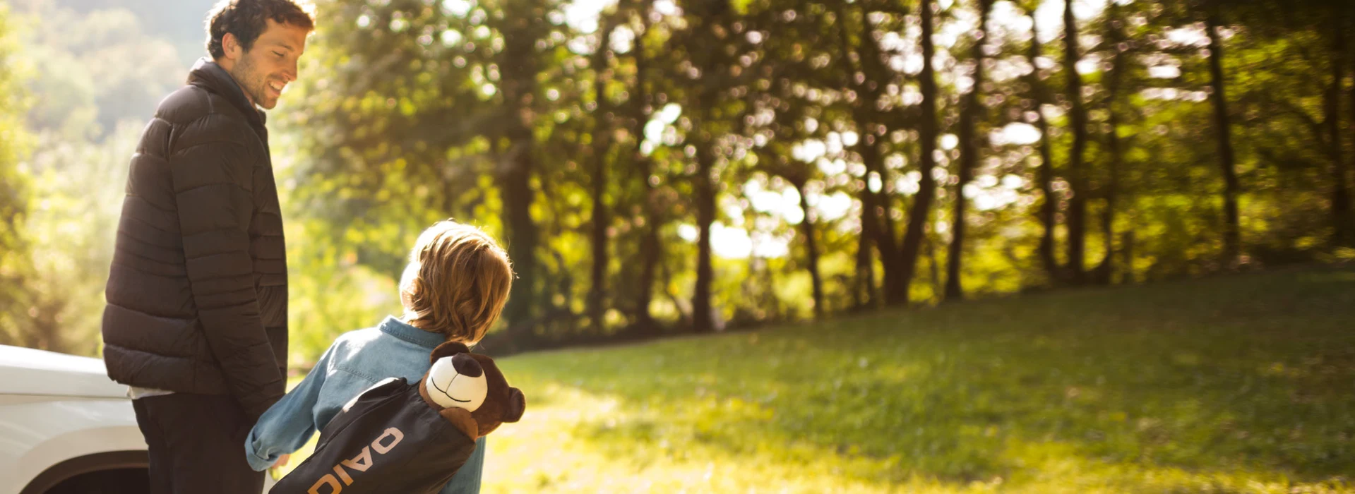 Vater und Kind mit Golfbag gehen bei Sonnenschein neben einem weißen Auto spazieren.