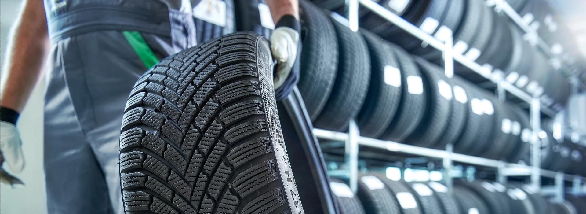 Eine Person mit Handschuhen hält einen Reifen in einem Lagerbereich, umgeben von gestapelten Reifen auf Regalen.