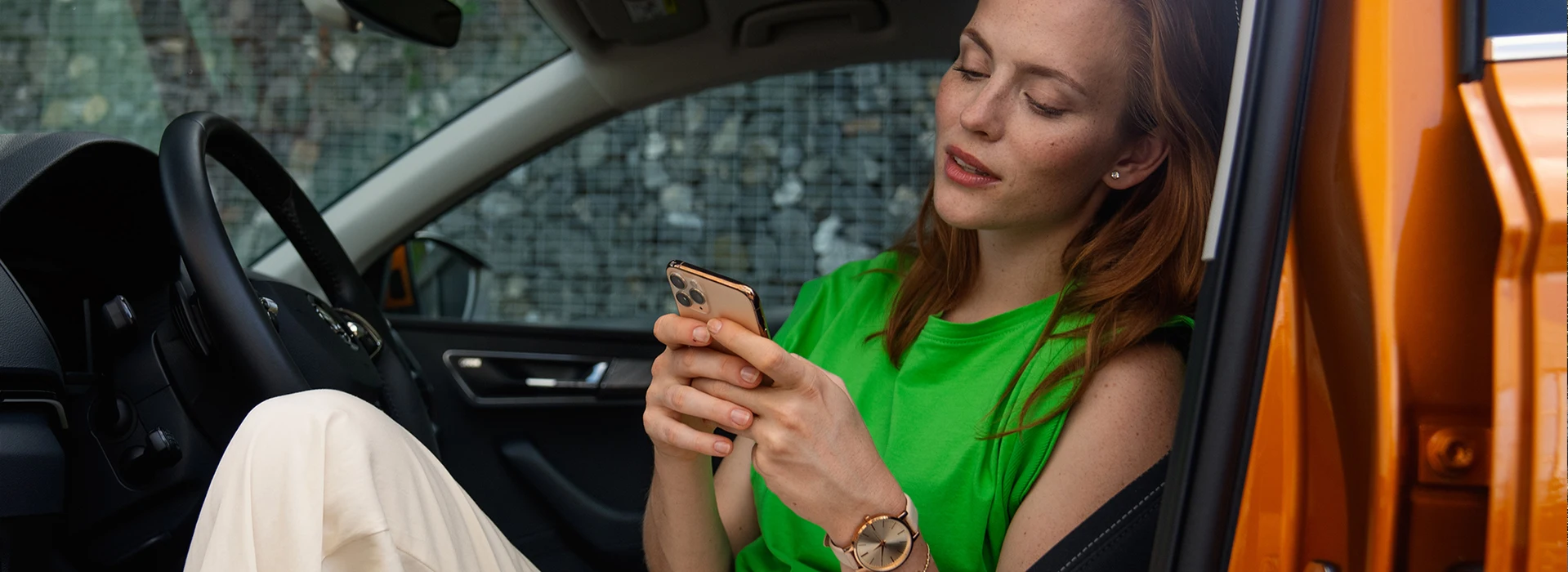 Eine junge Frau sitzt entspannt im Fahrerraum eines orangefarbenen Fahrzeugs, schaut auf ihr Smartphone und trägt eine grüne Weste, weiße Hose.