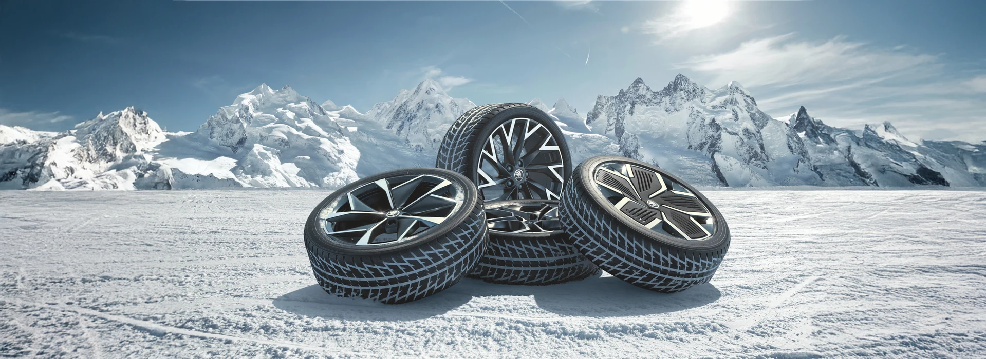 Drei Winterreifen mit markanten schwarzen Felgen liegen auf einer verschneiten Fläche vor einer bergigen Landschaft mit schneebedeckten Gipfeln. Der blaue Himmel mit Wolken und die sonnige Beleuchtung sind im Hintergrund zu sehen.