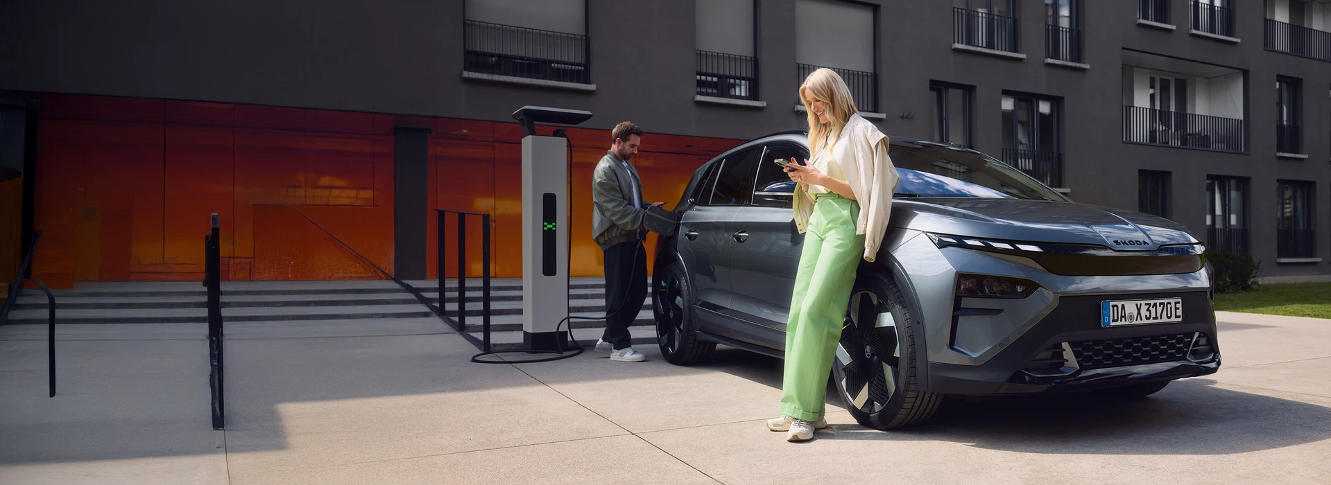 Zwei Personen laden ein Škoda Elektrofahrzeug an einer Ladesäule vor einem modernen Wohngebäude, während sie ihr Smartphone nutzen.