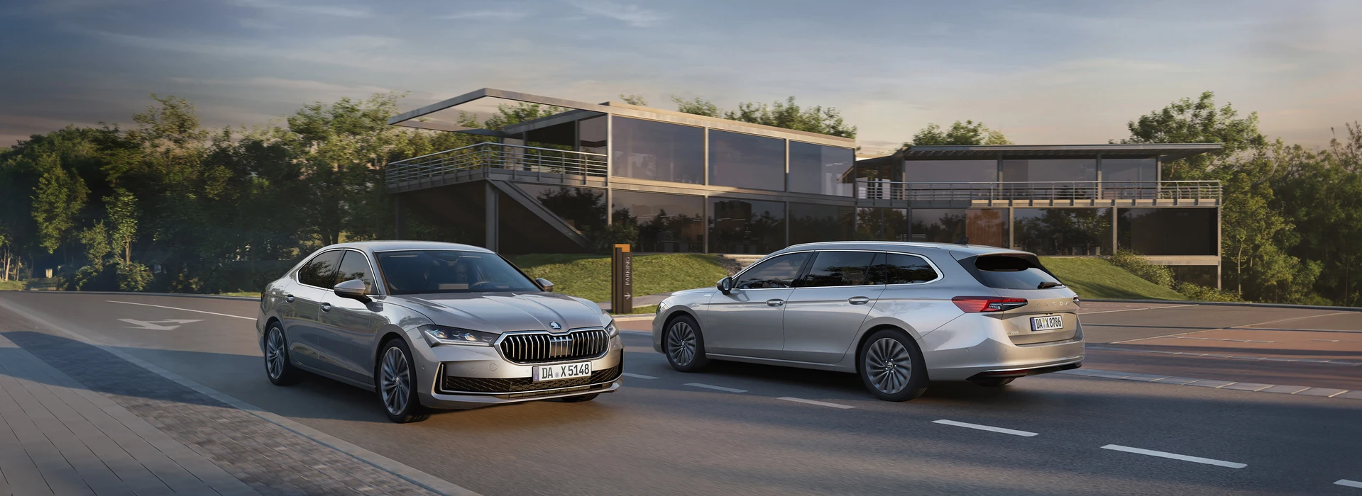 Zwei silberne Škoda Superb vor moderner Architektur – Limousine links, Kombi rechts – bei Sonnenuntergang auf der Straße.