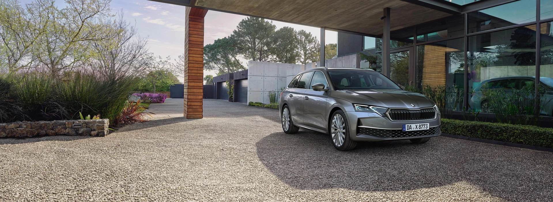 Frontansicht eines silbergrauen Škoda Octavia Combi vor einem modernen Haus mit Glasfassade, Garagen und Bäumen im Hintergrund.