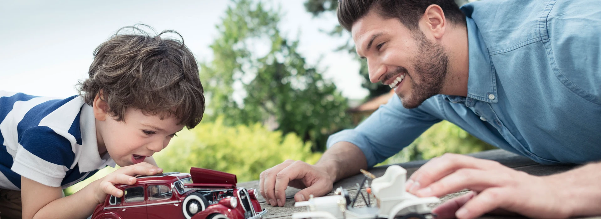Vater und Sohn spielen draußen mit Modellautos auf einem Tisch, beide lächeln