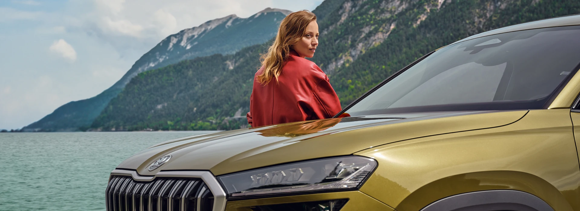 Frau lehnt entspannt an einem goldfarbenen Škoda Kodiaq, im Hintergrund ein See mit umliegenden Bergen.