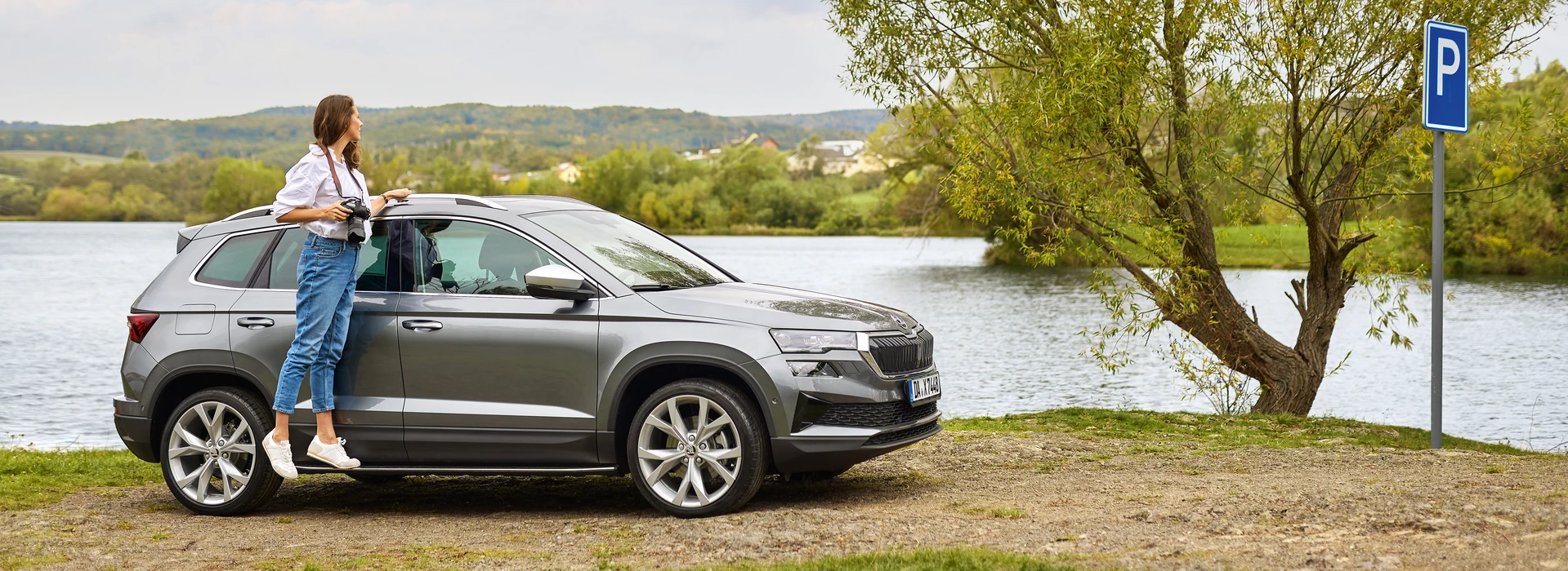 Frau lehnt mit Kamera am grauen Škoda Karoq, im Hintergrund ein See und ein P-Schild.