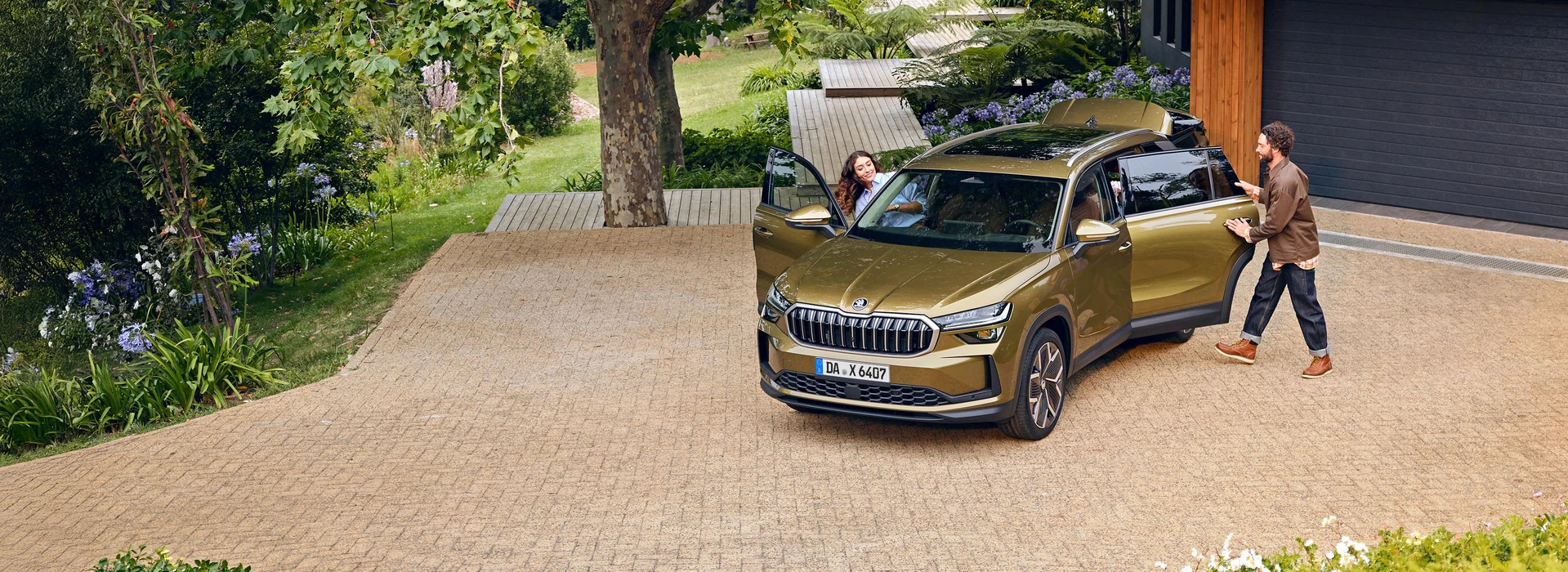 Ein Paar steht vor einem modernen Haus an einem goldfarbenen Škoda Kodiaq. Die Frau steigt auf den Beifahrersitz ein, der Mann öffnet die hintere Tür. Das SUV steht in einer gepflegten Einfahrt mit Blumen und Bäumen im Hintergrund.