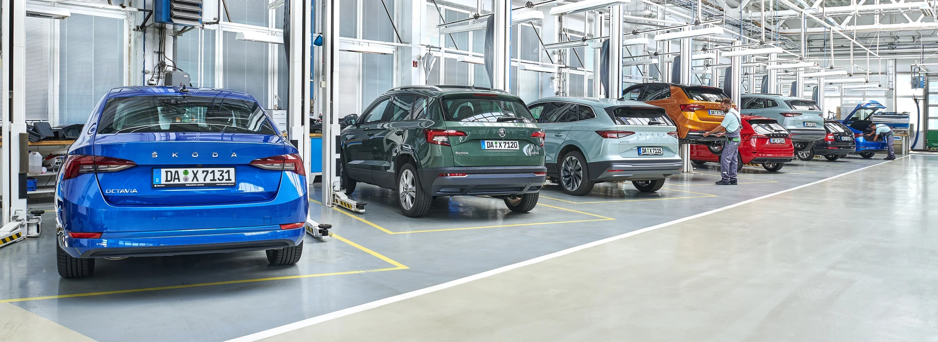 Sechs Škoda-Autos verschiedener Farben sind in einer gut beleuchteten Halle aufgereiht. Zwei Mitarbeiter führen Arbeiten an einem blauen und einem orangefarbenen Škoda-Fahrzeug durch. Verschiedene Maschinen und Geräte sind im Hintergrund sichtbar.