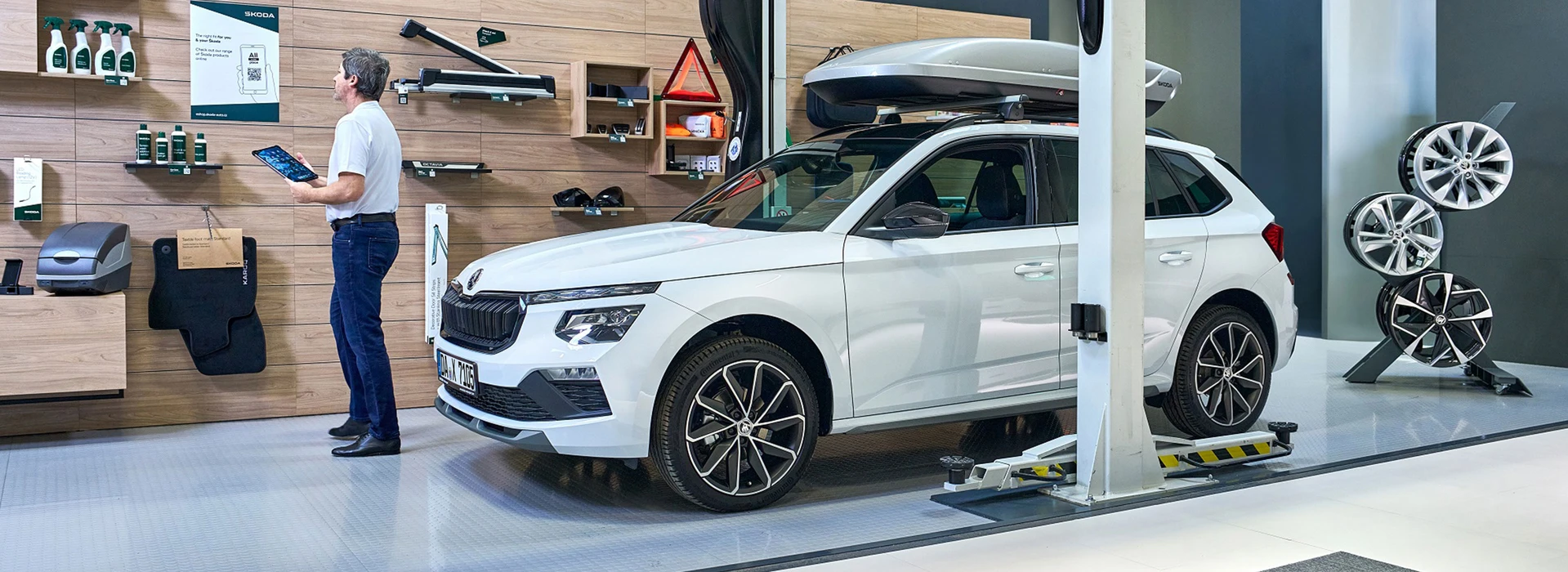 Weißer Škoda mit Dachbox steht in moderner Werkstatt, Mitarbeiter prüft Zubehör mit Tablet an Wand.