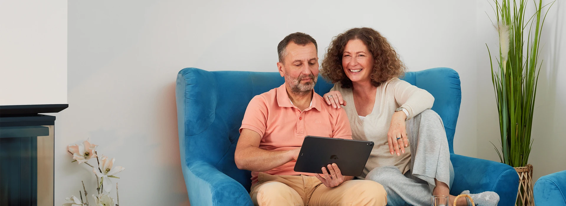 Paar sitzt entspannt auf blauem Sessel und schaut gemeinsam auf ein Tablet.