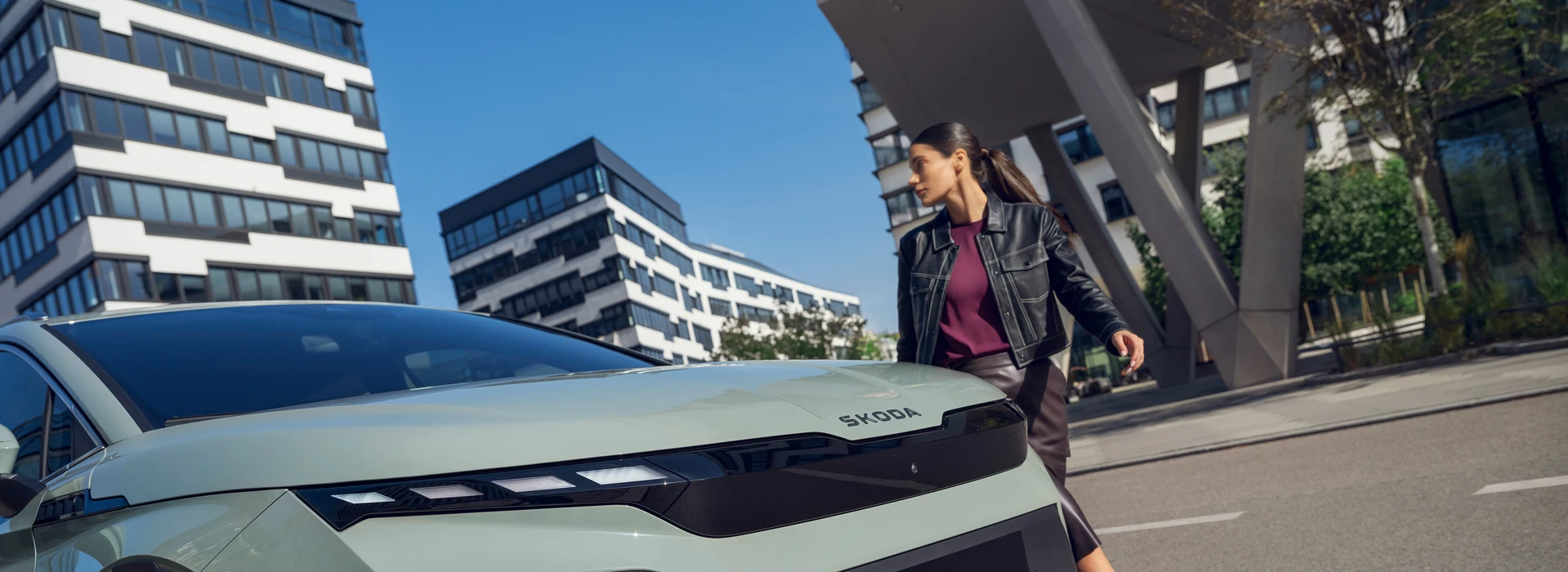 Eine Frau in schwarzer Lederjacke geht an einem hellgrünen Škoda Elroq vorbei, vor moderner Architektur.