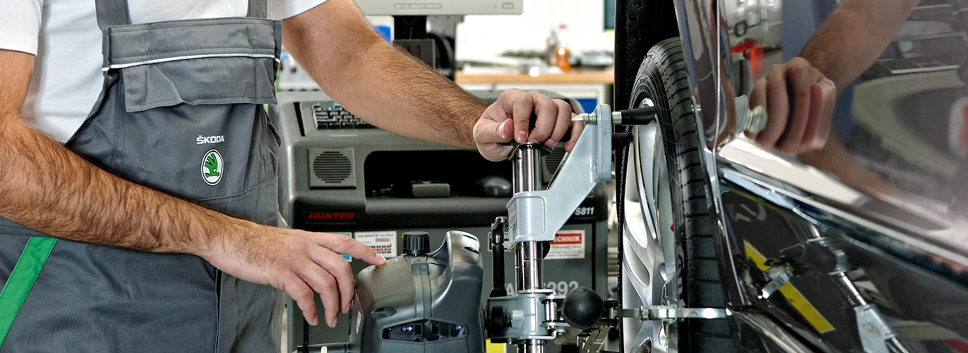 Ein Mechaniker in Škoda-Arbeitskleidung arbeitet an der Reifenauswuchtung eines Fahrzeugs in einer Werkstatt.