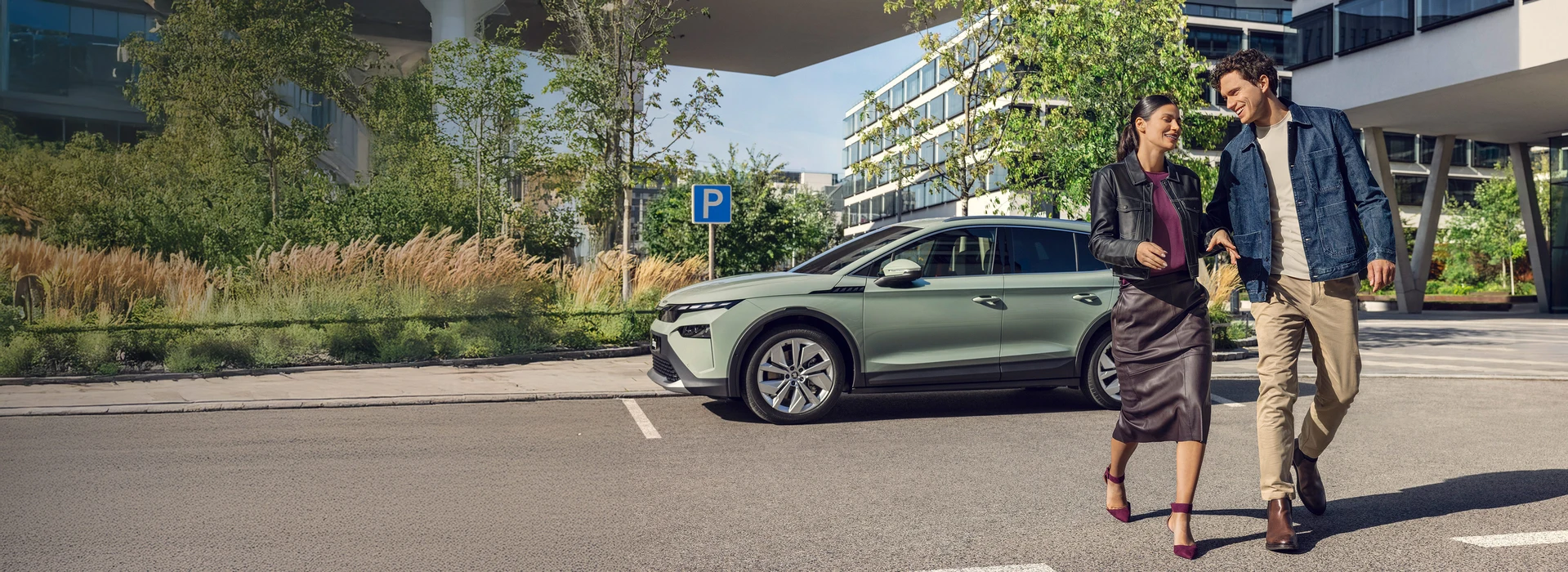Ein stilvoll gekleidetes Paar läuft lächelnd durch einen modernen, sonnigen Stadtbereich, im Hintergrund steht ein mintgrüner Škoda Enyaq auf einem Parkplatz. Die Umgebung zeigt moderne Bürogebäude, junge Bäume und einen blauen Parkplatz-Hinweis.